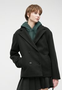 Black double-breasted coat with large buttons, notched lapels, and side pockets, layered over a green hooded sweatshirt and a pleated skirt.