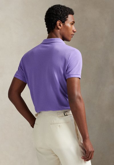 Mann in lavendelfarbenem Poloshirt und beiger Hose steht mit dem Rücken zu einem schlichten neutralen Hintergrund.