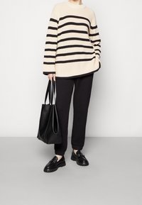 Kremowy sweter w czarne poziome paski, luźny krój, prążkowana faktura. Czarne spodnie dresowe, masywne czarne buty oraz czarna torba typu tote.