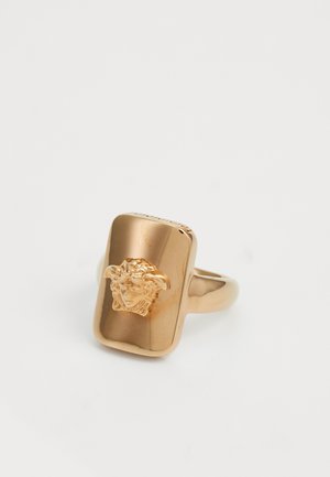 Bague en doré avec un design rectangulaire, dotée d'un détail sculptural en relief représentant un visage, texture lisse et finition polie.