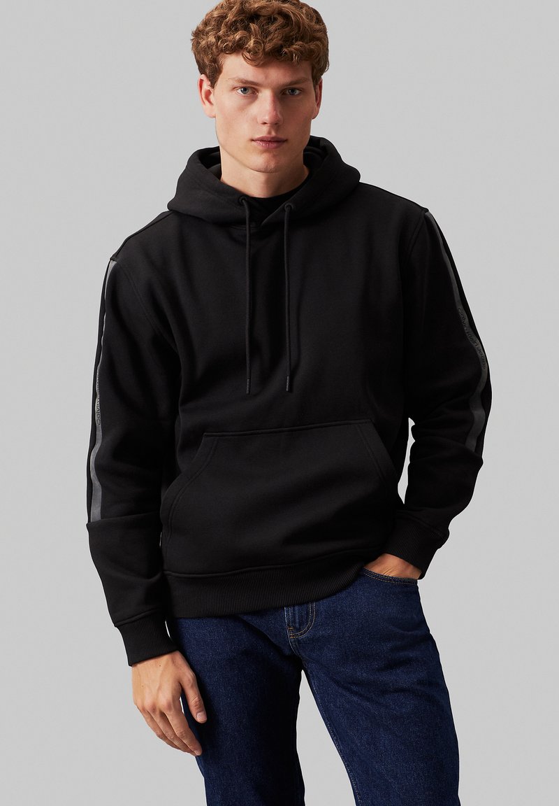 Felpa nera con cappuccio, tasca frontale, tessuto testurizzato, chiusure con coulisse e strisce laterali grigie a contrasto. Modello che indossa jeans di denim.