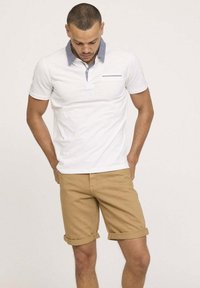 T-shirt polo blanc avec col bleu et poche poitrine, associé à des shorts en khaki retroussés. Texture de tissu lisse et design décontracté.