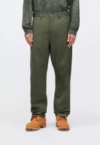 Zielone spodnie o prostych nogawkach wykonane z trwałego materiału, z przednimi kieszeniami, zapięciem na guzik i podwiniętymi mankietami. Dopasowane do beżowych butów.
