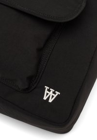 Musträpiga musta kott, millel on klapp, tikitud servade ja väikese valge tikandiga logo. Sile tekstuur ja ristkülikukujuline kuju.