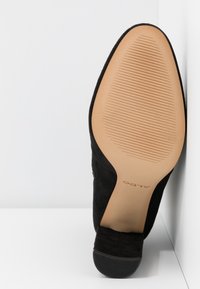 Chaussure à talon haut en daim noir avec une courbe lisse, un bout pointu et une semelle en caoutchouc beige avec des rainures horizontales pour l'adhérence.