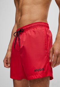Piros úszószort rövidnadrág könnyű anyagból, rugalmas derékpánttal, fekete húzózsinórral, és fekete "HUGO" logóval az alsó részen.