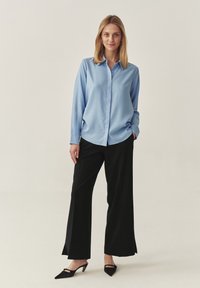 Cămașă lungă cu mâneci lungi de un albastru deschis, asortată cu pantaloni negri cu picioare largi și tocuri negre cu vârf ascuțit. Material neted și croială lejeră.