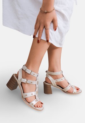 Femme portant des sandales blanches à talons avec brides à la cheville et clous, vernis rouge sur les doigts et les orteils, habillée d'un vêtement blanc.