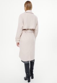 Jasno beżowy płaszcz z kołnierzem, wiązanym w talii i długimi rękawami. Projekt posiada skrócony tył. Osoba nosząca go zestawia go z czarnymi kozakami do kolan.