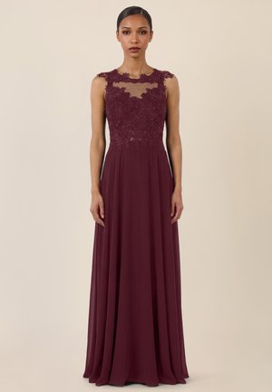 Frau trägt ein ärmelloses bodenlanges bordeauxrotes Kleid mit Spitze und Blumenstickerei am Oberteil sowie transparentem Ausschnitt.