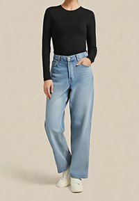 Osoba ubrana w czarną bluzkę z długim rękawem, jasnoniebieskie szerokie dżinsy z wysokim stanem oraz białe buty, stojąca na neutralnym tle.