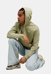 Sudadera verde con capucha, bolsillo canguro y logo en el pecho, combinada con jeans de pierna ancha en azul claro y zapatillas.