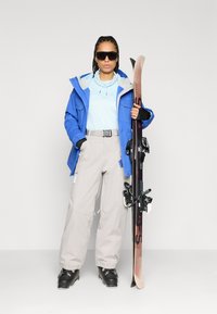 Lyžiarsky outfit zahŕňa modrú bundu so šedými akcentmi, svetlomodrý top, šedé nohavice a čierne lyžiarske topánky, pričom drží lyžu a slnečné okuliare.