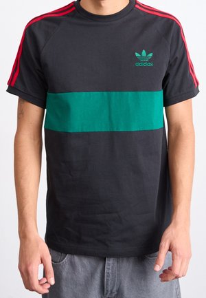 Homme portant un t-shirt Adidas noir avec des bandes rouges sur les épaules et une bande horizontale verte sur la poitrine.