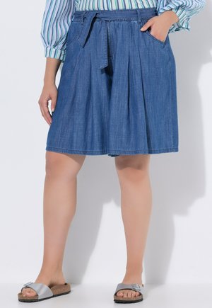 Kvinde iført blå knælange denimshorts med bælte, hænder i lommerne, stribet skjorte og sølvspænde-sandaler på hvid baggrund.