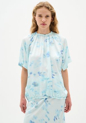 Lyseblå bluse med korte ærmer, rynket halsudskæring og elastisk kant. Har et subtilt blomstermønster i blå og lavendel nuancer.