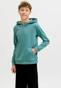 Teenagedreng med krøllet hår iført en grønblå hættetrøje med frontlomme og sorte bukser, smilende mod en ensartet lys baggrund.