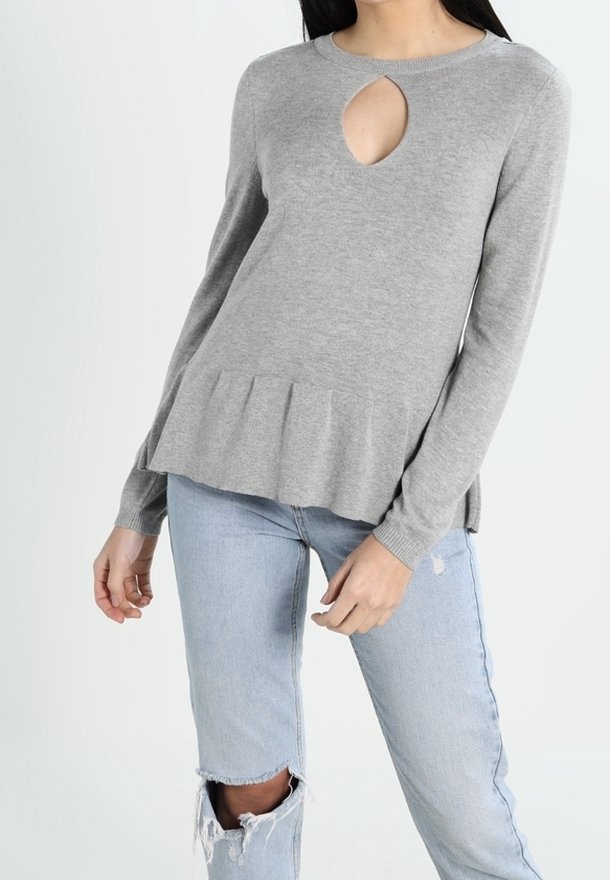 Femme portant un pull gris clair à manches longues avec une ouverture en forme de trou en clé devant et un ourlet à volants, associé à un jean bleu clair déchiré.