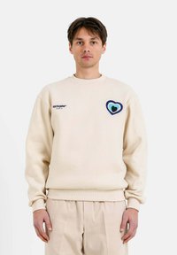 Sudadera beige con cuello redondo, que presenta un bordado de corazón multicolor en el lado izquierdo del pecho y puños y dobladillo acanalados. Corte informal.