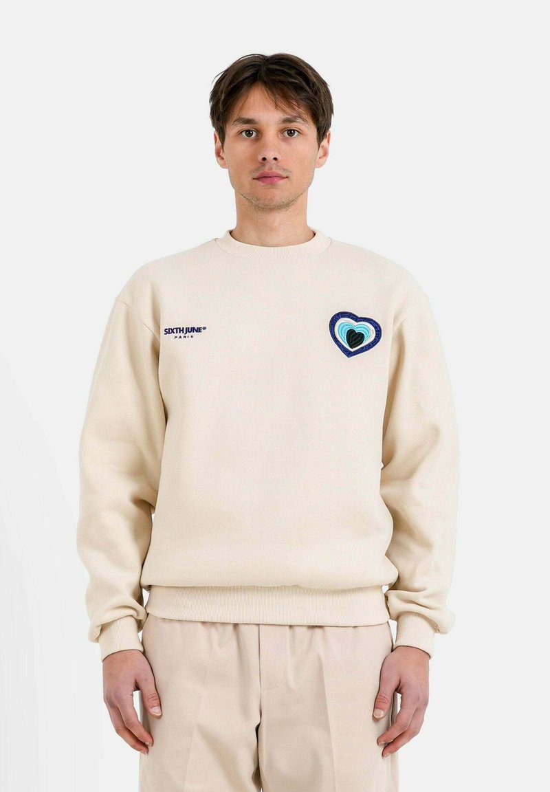Sudadera beige con cuello redondo, que presenta un bordado de corazón multicolor en el lado izquierdo del pecho y puños y dobladillo acanalados. Corte informal.