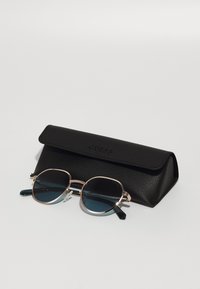 Occhiali da sole rotondi con montatura in oro metallico, lenti scure e custodia nera testurizzata etichettata "GUESS". La custodia ha una chiusura a pattina.