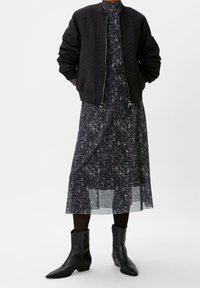 Schwarze gestepptes Bomberjacke mit gerippten Bündchen über einem schwarzen Kleid mit einem weißen abstrakten Muster. Schwarze Stiefeletten mit spitzen Zehen.