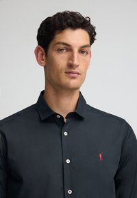 Camicetta nera con colletto strutturato, dotata di bottoni bianchi e di un piccolo logo rosso sul lato sinistro del petto. Tessuto dalla texture liscia.