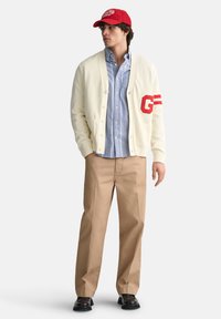 Cardigan crema con la "G" rossa e strisce, indossato sopra una camicia a righe blu chiaro, abbinato a pantaloni beige e un cappellino rosso. Scarpe nere.