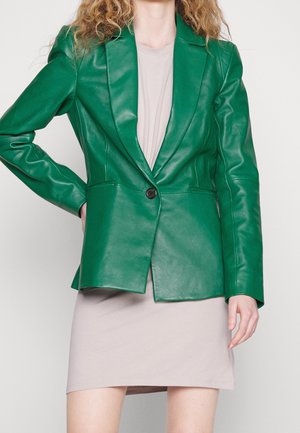 Persoon draagt een felgroene leren blazer met één knoop over een lichtbeige korte jurk tegen een effen achtergrond.