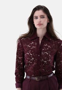 Burgunderfarbene Spitzenbluse mit floralen Mustern, einem Kragen und einer Knopfblende. Kombiniert mit hoch taillierten Hosen und einem passenden Gürtel.