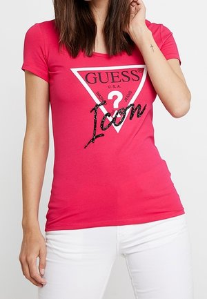 Kobieta nosząca dopasowany, jaskraworóżowy t-shirt Guess z białym trójkątnym logo i czarnym napisem "Icon", zestawiony z białymi spodniami.