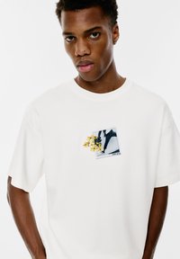 Weißes Baumwoll-T-Shirt mit einem vorderen grafischen Design eines Fotos, das mit gelber und grüner floralem Stickerei verziert ist. Kurze Ärmel, lockere Passform.