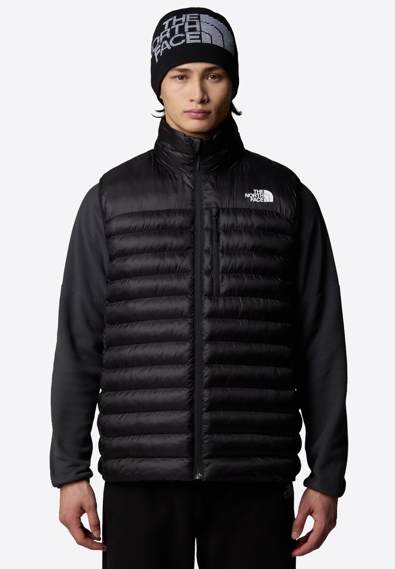 Zwarte puffer bodywarmer met horizontale baffles en een volledige ritssluiting. Gecombineerd met een zwarte lange mouwen top en een gebreide muts met het merklogo.