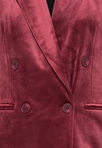 Burgundská sametová sako s dvojitým zapínáním, které má dva kulaté knoflíky a boční kapsy. Měkká textura s hladkým povrchem.