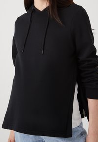 Black hoodie made from a smooth material with a V-neck, long sleeves, and side slits. Features adjustable drawstrings and a layered look.