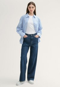 Camisa de rayas azul y blanca de gran tamaño, camiseta blanca, jeans oscuros de cintura alta y zapatillas blancas en una pose de pie.