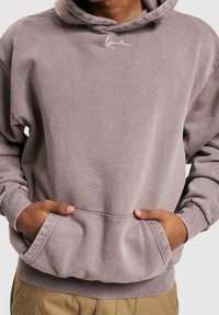 Persona con una sudadera mauve desgastada y un pequeño logotipo blanco bordado, con las manos en el bolsillo delantero, combinada con pantalones marrón claro.