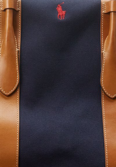Bolsa de lona azul-marinho com detalhes em couro castanho, apresentando um pequeno logótipo de polo bordado em vermelho. Costura decorativa ao longo das bordas.