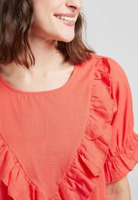 Chemise corail avec un décolleté dégagé et des volants bien marqués sur le devant et les manches ; fabriquée en tissu léger.