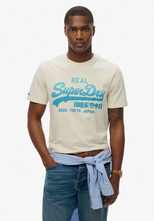 T-shirt crema con testo grafico blu "REAL SuperDry" e caratteri giapponesi. Abbinato a jeans blu e camicia azzurra chiara annodata in vita.