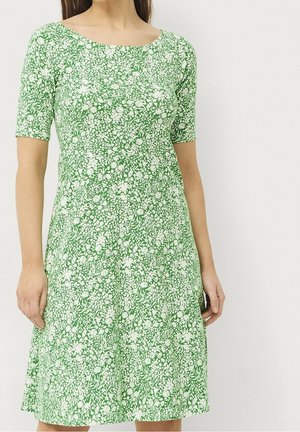 Femme portant une robe verte mi-longue à manches courtes avec un motif floral blanc, debout sur un fond clair uni.