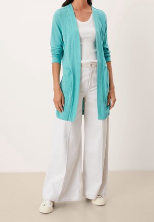 Nainen, jolla on päällään vaaleanturkoosi avonainen neuletakki valkoisen topin, valkoisten leveälahkeisten housujen ja valkoisten tennareiden päällä.