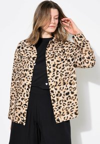 Hedvábná košile s leopardím potiskem v béžové a černé barvě, s dvěma náprsními kapsami a volným střihem, nošená přes černý top a kalhoty s širokými nohavicemi.