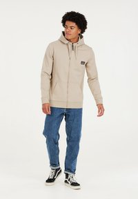 Beige Zip-Hoodie mit einer Fronttasche, Kordelzugkapuze und schwarzem Logo-Patch, kombiniert mit blauen, schmal geschnittenen Jeans und schwarzen Turnschuhen.