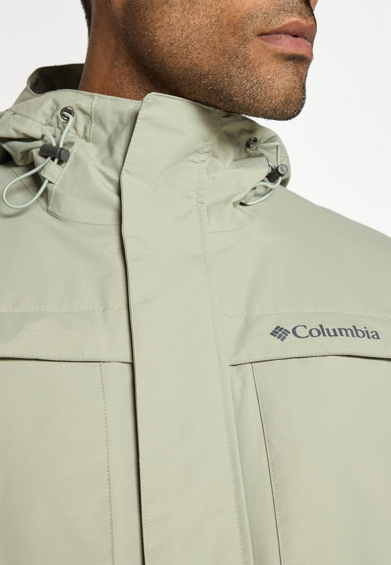 Jasnozielona kurtka Columbia z wysokim kołnierzem, regulowanymi sznurkami i dwiema przednimi kieszeniami. Posiada gładki, teksturowany materiał oraz detal z logo.