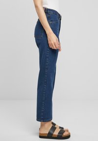 Jeans de mezclilla azul de cintura alta con un corte recto, con cierre de botón y dos bolsillos traseros. Combinados con sandalias negras con tiras.