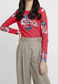 Punainen pitkähihaine toppi sinisillä ja vaaleanpunaisilla kukkakuoseilla, yhdistettynä korkeavyötäröisiin beigeihin housuihin, jotka on valmistettu tekstuuripinnoitteisesta kankaasta.