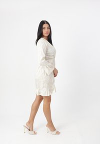 Cremfarbendes Blumenkleid mit langen Ärmeln und rüschengesäumtem Saum; kombiniert mit beigen, hochhackigen Mules mit einem Kreuzgurt-Design.