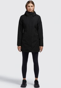 Zwarte waterdichte parka met capuchon, twee met rits afgesloten zijzakken en een verlengd ontwerp. Draag over zwarte legging en sneakers. Glad van structuur.