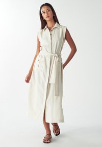 Donna che indossa un vestito senza maniche beige con bottoni e cintura, dotato di ampie tasche, abbinato a sandali neri con cinturini, in piedi su uno sfondo bianco.